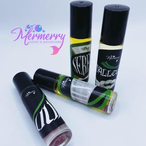May include: Four glass roller bottles with black caps and labels. The labels are black with white and green text. The text on the labels reads "Mermerry Soaps & Bathbombs", "Lily", "Valley", and "Serene".