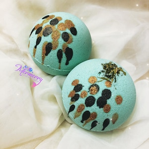May include: Two round, light blue bath bombs with gold and black decorative accents. One bomb has dried herbs on top. The bath bombs are on a white, textured surface. The logo "Mermerry Soaps & Bath Treats" is in the lower left corner.