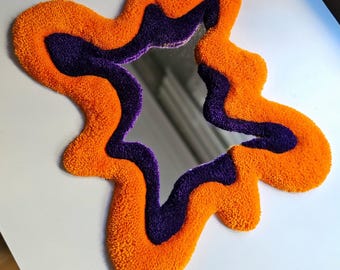 Può includere: Specchio astratto con un bordo morbido e strutturato in arancione e viola. La forma dello specchio è irregolare, con un tocco morbido e tattile. Il bordo è arancione brillante, con un interno viola scuro. Un moderno elemento decorativo.