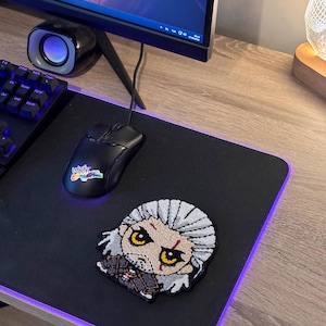 Könnte beinhalten: Schwarze Gaming-Maus und -Mauspad mit lila LED-Kante. Das Mauspad zeigt einen Cartoon-Patch eines Charakters mit weißen Haaren und gelben Augen. Ein Computermonitor, eine Tastatur und ein Lautsprecher befinden sich im Hintergrund.