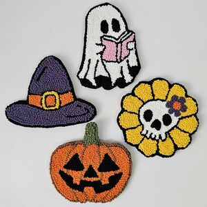 May include: A collection of handmade Halloween-themed decorative items. Includes a purple witch hat with an orange buckle, a white ghost reading a pink book, a jack-o'-lantern, and a skull-shaped flower with yellow petals.