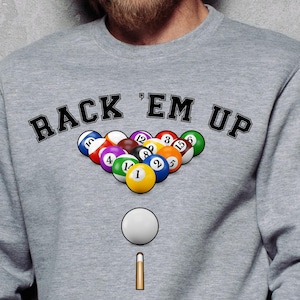 Op de afbeelding: Een grijze sweatshirt met de tekst "BACK 'EM UP" in zwarte letters. Daaronder een afbeelding van biljartballen, een witte bal en een keu. De biljartballen zijn in een driehoek gerangschikt.