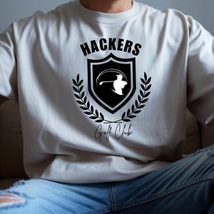 Könnte beinhalten: Hellbeige T-Shirt mit einem schwarz-weißen Golf-Motiv. Das Motiv zeigt einen Golfspieler beim Abschlag in einem Schild, mit dem Schriftzug "Hackers Golf Club" in Schwarz. Lorbeerblätter umgeben das Design.