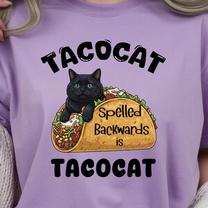 May include: A lilac t-shirt with a black cat inside a taco. The text above reads "TACOCAT", and below the taco, it says "Spelled Backwards is TACOCAT". The taco is filled with vegetables and meat.