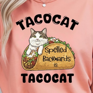 May include: Peach-coloured t-shirt with a graphic of a cat in a taco. The text above reads "TACOCAT", with "Spelled Backwards is TACOCAT" below the taco. The cat has white and grey fur and green eyes.