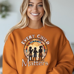 May include: An orange sweatshirt featuring the words "Every Child Matters" in a circular design. The design includes silhouettes of three children. The sweatshirt is a solid colour and has long sleeves.