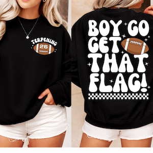 Può includere: Felpa nera con la scritta "BOY GO GET THAT FLAG!" sul retro. La parte anteriore presenta una grafica di football con la scritta "TERPENING 26". Il design è in bianco e marrone.