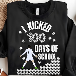 以下が含まれることがあります： サッカーボールを蹴るサッカー選手の白いグラフィックが描かれた黒いスウェットシャツ。グラフィックには「100日間の学校を蹴った」と書かれています。
