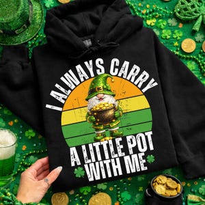 May include: Black hoodie with a graphic of a leprechaun holding a pot of gold, set against a green, yellow, and orange background. The text reads "I ALWAYS CARRY A LITTLE POT WITH ME" with shamrocks. A festive St. Patrick's Day theme.