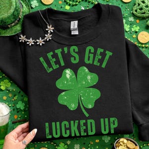 Peut inclure: Sweat-shirt noir avec l'inscription verte "LET'S GET LUCED UP" et un trèfle à quatre feuilles vert. Un chapeau vert de la Saint-Patrick et des pièces d'or sont en arrière-plan. Un collier argenté avec des breloques en forme de fleurs est sur le sweat-shirt.