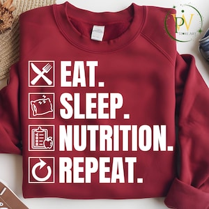 May include: A maroon sweatshirt with white text and icons. The text reads "EAT. SLEEP. NUTRITION. REPEAT." with corresponding icons of a knife and fork, a pillow, a clipboard with an apple, and a recycling symbol. The sweatshirt is folded.