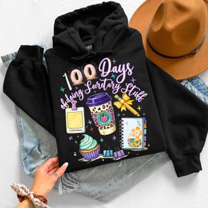 May include: Black hooded sweatshirt with a colorful graphic. The graphic includes the text "100 Days of doing Secretary Stuff", a coffee cup, a cupcake, a notepad, and other office-themed illustrations.