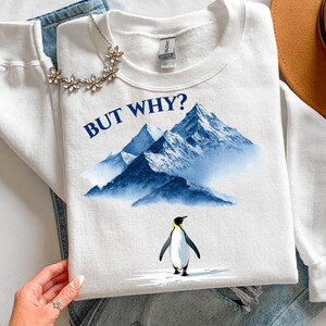 Könnte beinhalten: Weißes Sweatshirt mit dem Text "BUT WHY?" über einer blauen Berglandschaft und einem Pinguin. Der Pinguin hat einen schwarzen Körper, einen weißen Bauch und gelbe Akzente. Eine Blumenkette ist sichtbar.