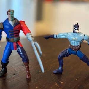 May include: Two action figures of DC Comics characters, Two-Face and Batman, are displayed. Two-Face is in a blue and red outfit, holding a weapon. Batman is in a light blue and dark blue costume, with a bat symbol on his chest.