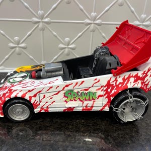 May include: A white toy car with red flame graphics and the word Spawn printed on the side. The vehicle features a silver engine, a black interior with chain details, silver wheels wrapped in chains, and a large red spoiler in the upright position.