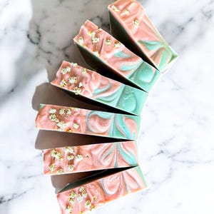 Puede incluir: Barras de jabón artesanales con un diseño en espiral rosa, turquesa y blanco. Cada barra está adornada con copos de oro y pequeñas flores blancas. Los jabones están dispuestos en forma de abanico sobre una superficie de mármol blanco.
