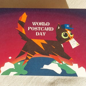 May include: A rectangular postcard featuring a stylised black and orange cat wearing a blue mail carrier hat, holding a white postcard in its mouth. The cat stands on a globe against a purple and red gradient background with the text WORLD POSTCARD DAY.