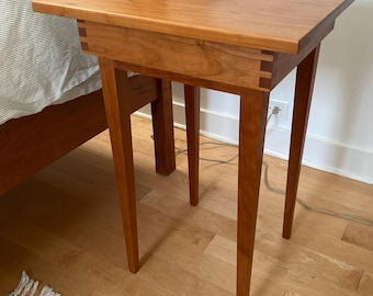 May include: A small wooden end table with a cherry wood finish and four legs. The table has a simple design and is perfect for a bedroom or living room.