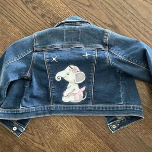 May include: A blue denim jacket with a cute embroidered elephant wearing a pink tutu and a flower on its head. The elephant is on the back of the jacket.