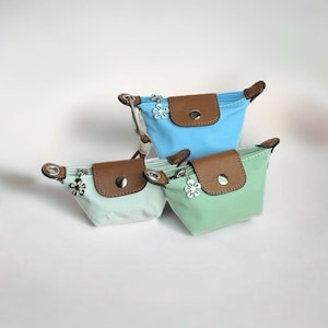 Puede incluir: Tres bolsos pequeños en azul claro, verde pálido y verde menta. Cada bolso cuenta con una solapa marrón texturizada con un botón a presión plateado, una cremallera plateada y un pequeño dije plateado en forma de flor.