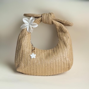 May include: A crescent-shaped handbag crafted from woven tan straw material. The bag features a knotted top handle and is decorated with a large, white, five-petaled flower accent on the left side, along with a small white flower charm.