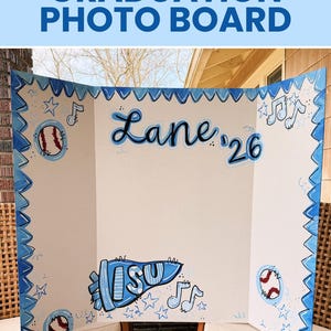 May include: A white tri-fold graduation photo board featuring hand-painted blue accents. The board displays the name Lane, the year '26, a blue pennant with the letters ISU, baseballs, musical notes, and stars, all framed by a blue zigzag border.