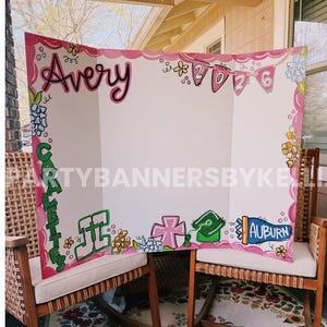 May include: A white tri-fold display board featuring hand-painted pink scalloped borders, floral accents, and the name Avery in black script. It includes the year 2026 on pink pennants, the word CARALETTES in green, and the text AUBURN in blue.