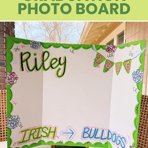 May include: A white tri-fold graduation photo board with a green scalloped border. The board features the hand-lettered name Riley, a banner with the year 2026, and the text IRISH -> BULLDOGS. The design includes floral accents in pink and blue.