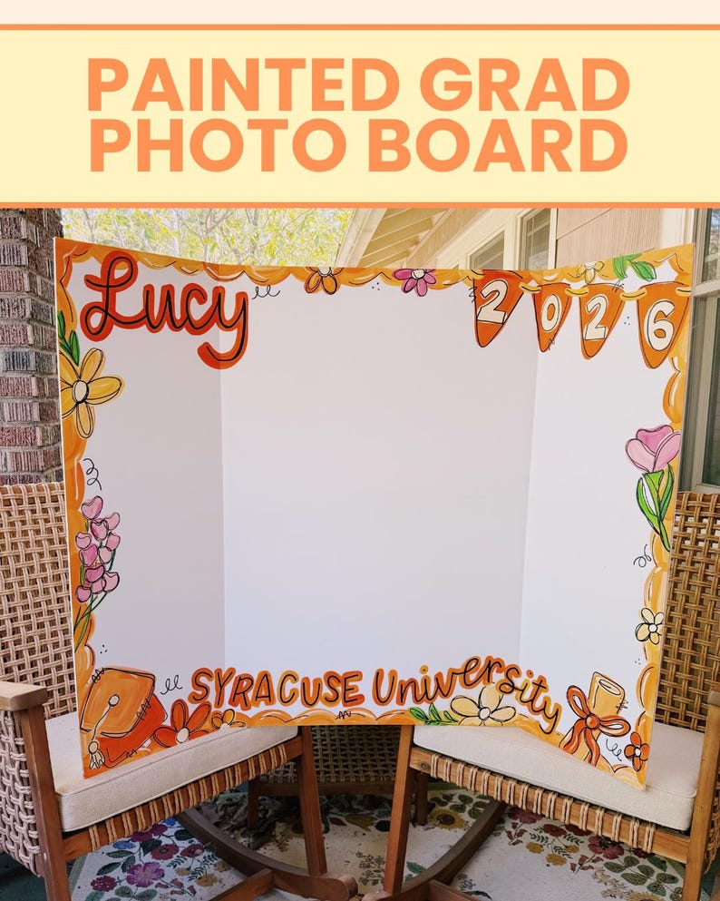 May include: A white tri-fold display board featuring hand-painted orange and yellow floral borders. The board displays the name Lucy, the year 2026 on orange pennants, and the text Syracuse University, along with a painted graduation cap and diploma.