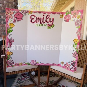 May include: A white tri-fold display board featuring a pink scalloped border decorated with hand-drawn pink, yellow, and orange flowers. The text reads Emily Class of 2026 in black and green lettering, with a pink graduation cap illustration.