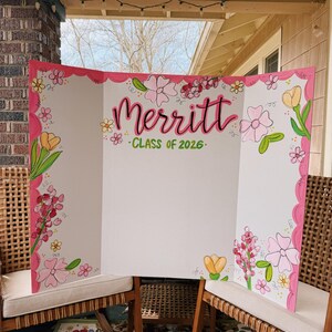 May include: A white tri-fold display board featuring a hand-painted floral border in shades of pink, yellow, and green. The centre displays the name Merritt in large pink script lettering above the green text "CLASS OF 2026" on a white background.