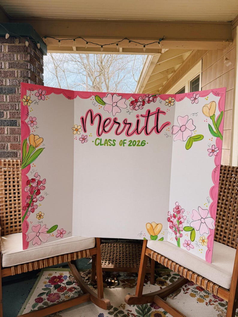 May include: A white tri-fold display board featuring a hand-painted floral border in shades of pink, yellow, and green. The centre displays the name Merritt in large pink script lettering above the green text "CLASS OF 2026" on a white background.