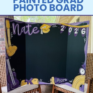 May include: A tri-fold black display board featuring hand-painted purple and yellow graduation-themed illustrations. The board displays the name Nate, the year 2026, a graduation cap, a guitar, a diploma, and the text Lipscomb in yellow paint.