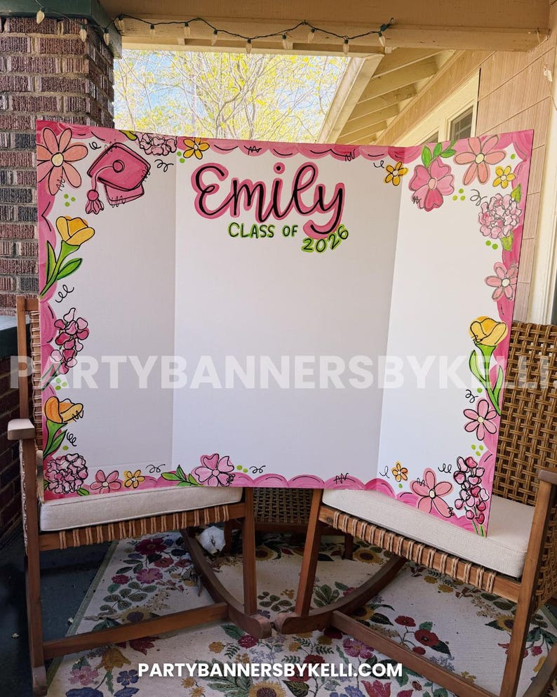 May include: A white tri-fold display board featuring a pink scalloped border decorated with hand-drawn pink, yellow, and orange flowers. The text reads Emily Class of 2026 in black and green lettering, with a pink graduation cap illustration.