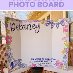 May include: A tri-fold white display board featuring hand-painted floral borders in pink, yellow, and purple. The board displays the name Delaney, the year 2026 on a banner, and text reading TCU Honor's College and Strategic Communications & Business Analytics Class of 2030.