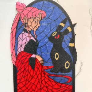 May include: A stained-glass style ornament featuring a woman with pink hair and a black Umbreon. The woman wears a black dress and red robe. The background is blue with black lines, and the ornament has a black border and a loop for hanging.