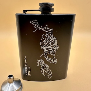 Könnte beinhalten: Schwarze Edelstahl-Taschenflasche mit einer weißen Umrisskarte von Stowe, Vermont, und den Koordinaten 44.5305° N - 72.7803° W. Die Flasche hat einen schwarzen Verschluss und wird mit einem silbernen Trichter geliefert.