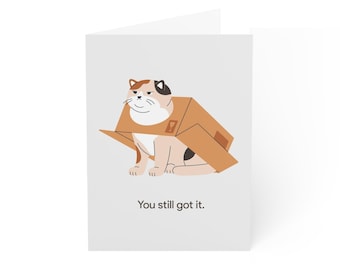 May include: A folded greeting card with a cartoon cat inside a cardboard box. The cat is depicted in tan, brown, and black. The text "You still got it." is printed below the image, which is set against a white background.