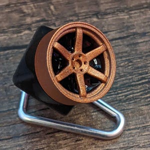 May include: A bronze-colored miniature car wheel with six spokes, mounted on a black base. The wheel is set on a silver metal stand. The wheel's design is detailed, and the overall aesthetic is automotive-themed.