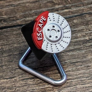 Op de afbeelding: Een zilverkleurige remschijf met een rode remklauw met de tekst "ESCAPE" is gemonteerd op een zwarte standaard met een zilverkleurige metalen basis. De remschijf heeft meerdere gaten langs de rand. De achtergrond is een houten oppervlak.