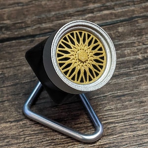 May include: A miniature gold-colored wheel rim model with a silver outer ring and a black base. The wheel is supported by a silver metal stand. The design features a detailed spoke pattern, showcasing a car accessory.