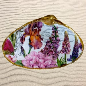 May include: A decorative seashell with a gold-painted rim, featuring a floral design with purple, pink, and orange flowers. The interior has a white background with script text. The shell is resting on a sandy surface.