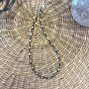 May include: A silver necklace with oval-shaped beads. The necklace is laying on a woven straw surface.