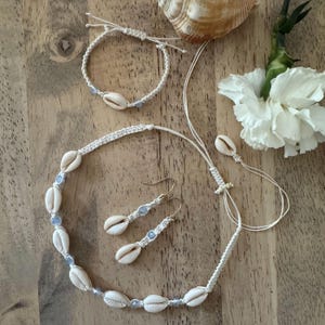 May include: A handmade jewelry set featuring a white macrame necklace, bracelet, and earrings, all adorned with seashells and light blue beads. The set is displayed on a wooden surface, with a seashell and white flower in the background.