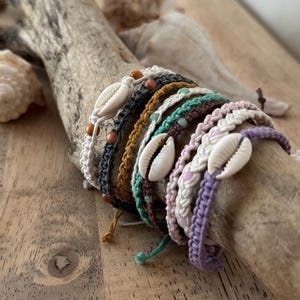 May include: A collection of handmade macrame bracelets in various colors, including white, gray, tan, teal, pink, and purple. Each bracelet features a cowrie shell and wooden or glass beads. The bracelets are displayed on a piece of driftwood.