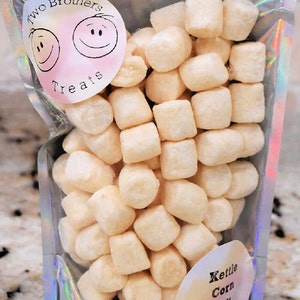May include: A clear plastic bag filled with small, white, kettle corn soft puffs. The bag has a label that says "Two Brothers Treats" with two smiling faces. Another label says "Kettle Corn Soft Puffs".
