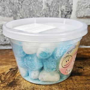 May include: A clear plastic container filled with blue and white candy. The candy is shaped like hearts and is covered in a white sugar coating. The container has a sticker on the side that says "Two Bros Treats".