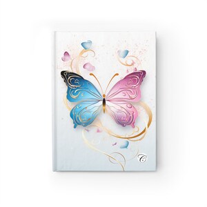 Puede incluir: Un diario de tapa dura con una cubierta blanca que presenta una ilustración central de una mariposa. La mariposa tiene el ala izquierda azul y el ala derecha rosa con patrones de filigrana dorados, espirales doradas y un logotipo de la letra C.