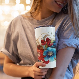 Puede incluir: Un vaso cilíndrico blanco con un diseño floral vibrante que presenta grandes flores rojas y azul brillante, acentuado por intrincadas líneas doradas en espiral y flores más pequeñas de color azul claro, con una tapa de plástico transparente.