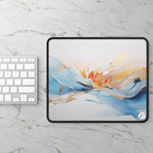 Puede incluir: Alfombrilla de ratón rectangular con borde cosido negro. La superficie presenta un diseño abstracto con pinceladas fluidas azules, naranjas y doradas sobre un fondo blanco. Se ve un teclado blanco.